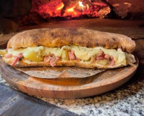 Panuozzo Montagnard: Pain cuit au feu de bois, farine BIO. Sandwich base crème fraîche BIO de Normandie, lardons Label Rouge, pommes de terre et oignons cuits au four. La touche final, notre raclette artisanale AOP fondue dans notre four au feu de bois.